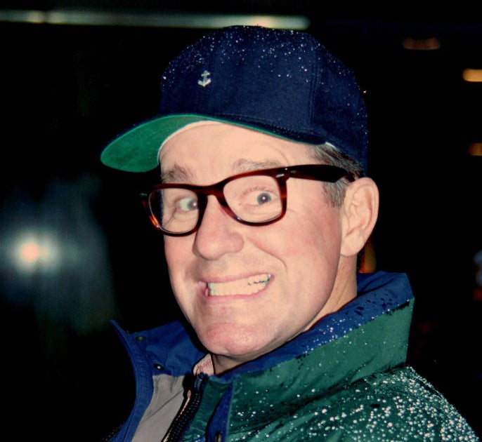 Phil Hartman in glasses making a surprised expression wears a cap and a jacket with raindrops