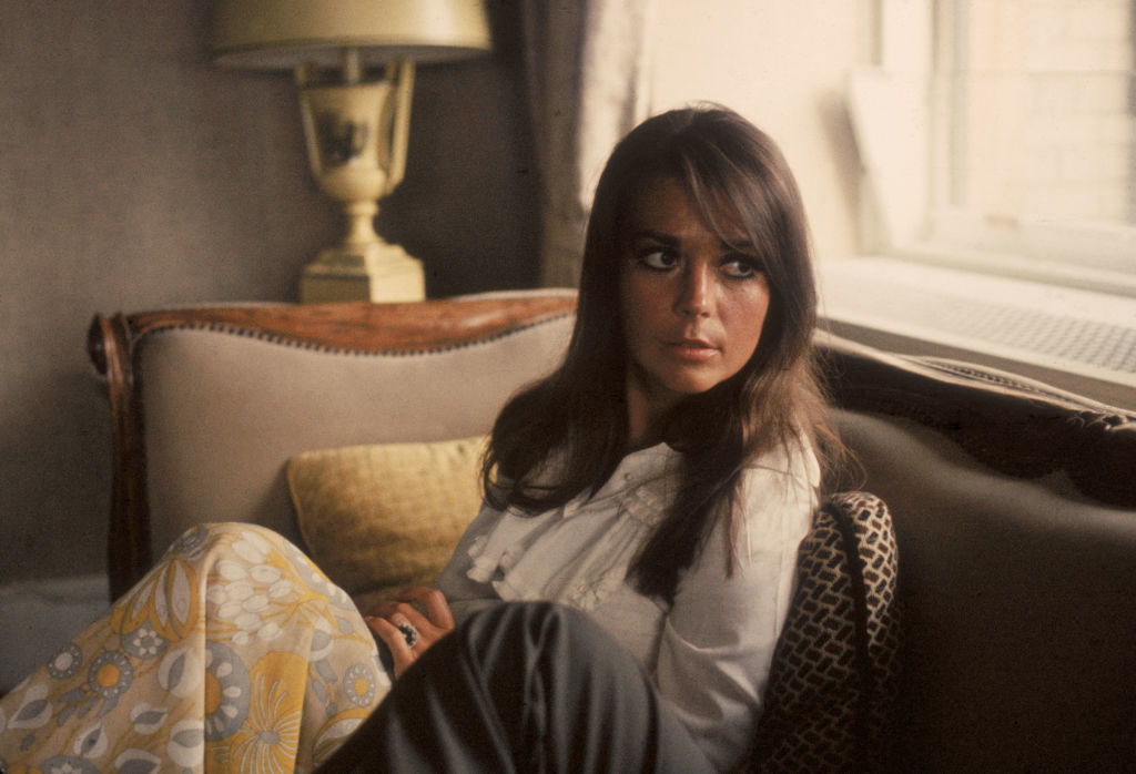 Wood seated on a couch, looking pensive. She wears a patterned outfit