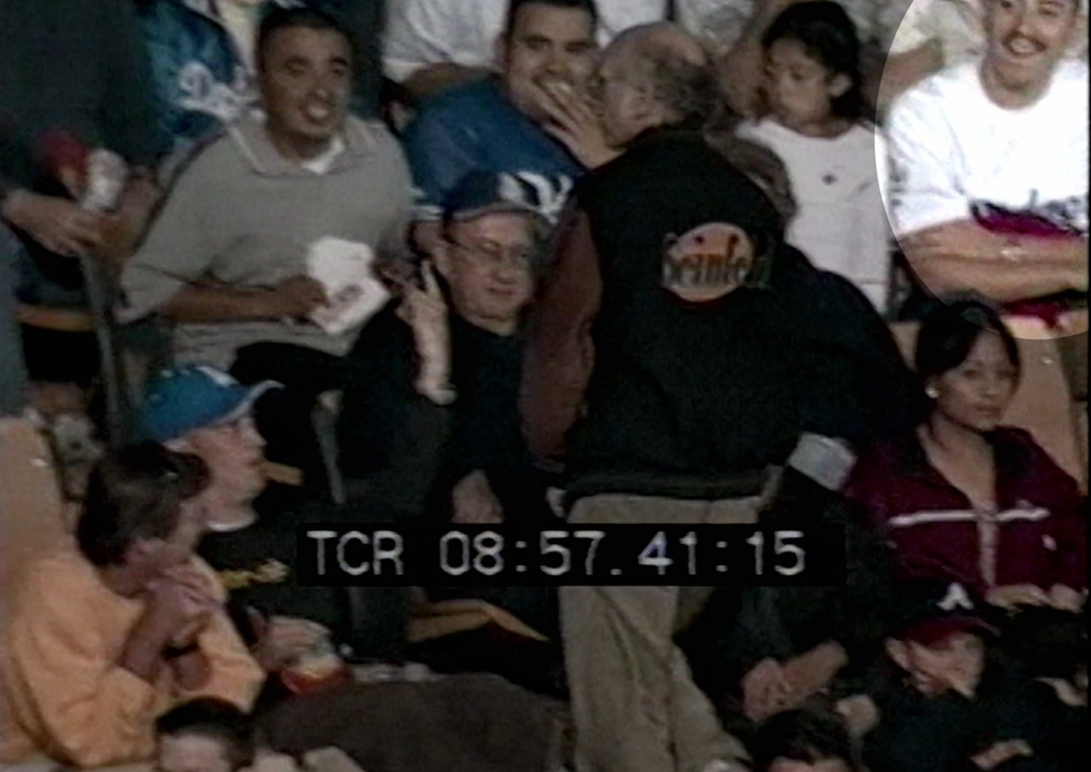 A person stands facing a crowd where some audience members are smiling and looking towards the camera; timecode overlay is present