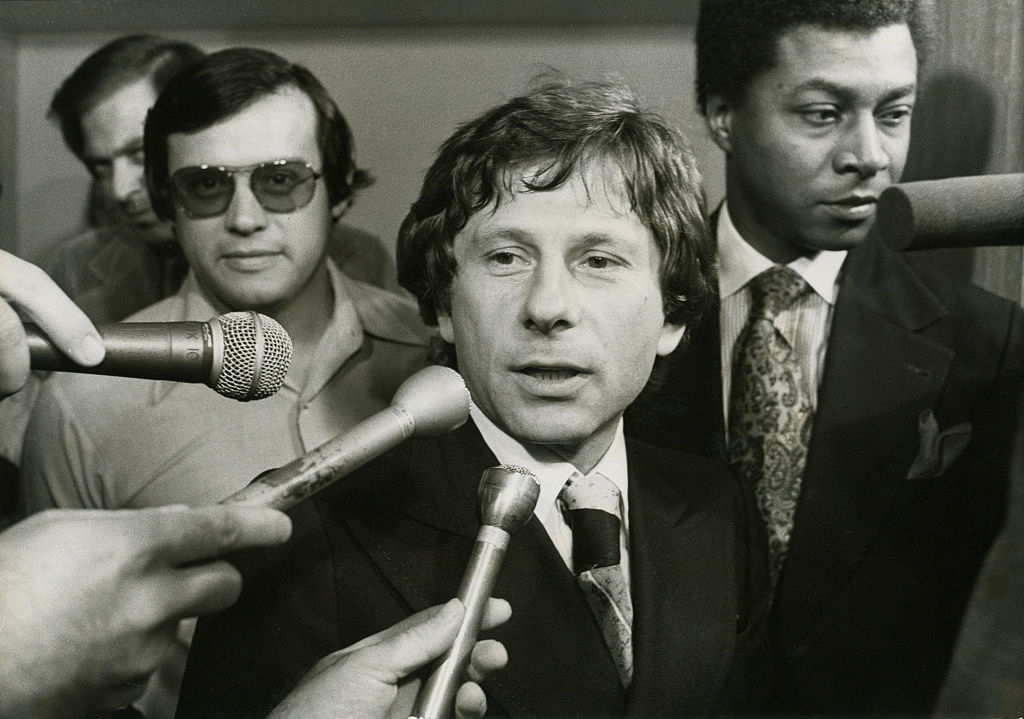 Polanski surrounded by reporters holding microphones, speaking at an event