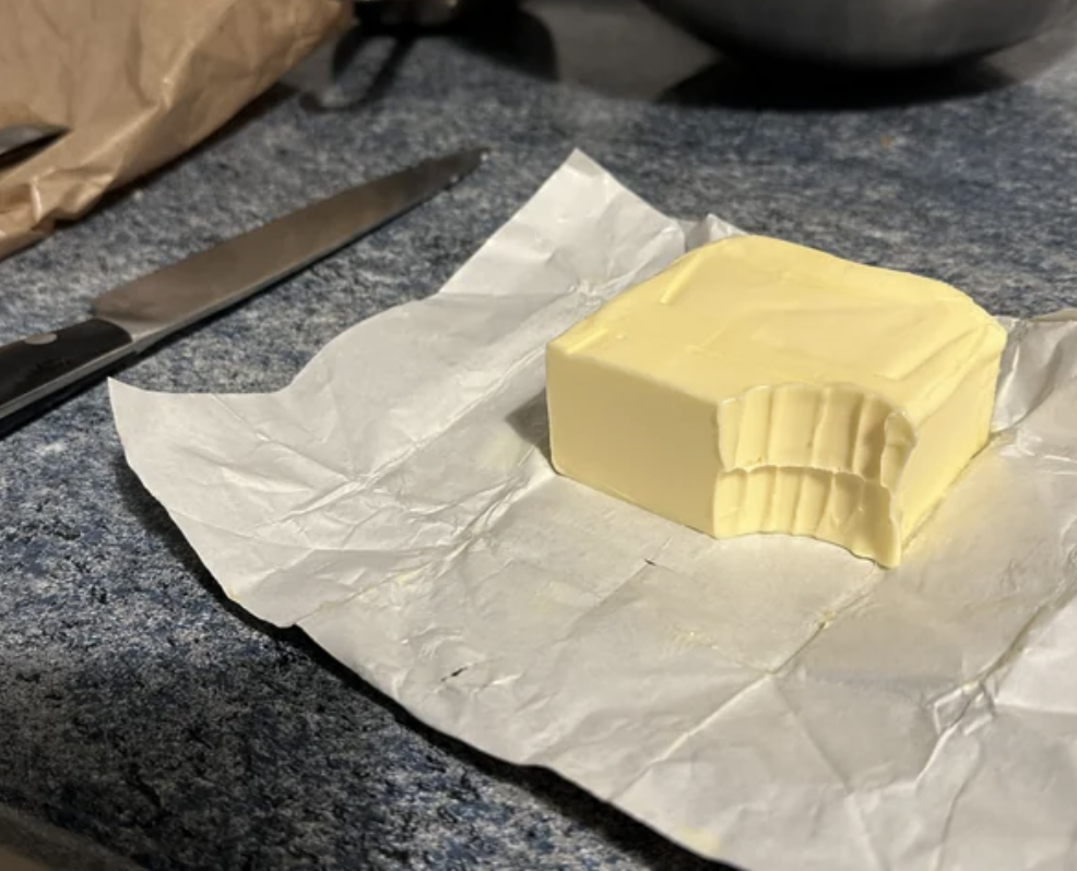 A stick of butter with a few slices cut off, sitting on its wrapper, next to a knife on a kitchen counter
