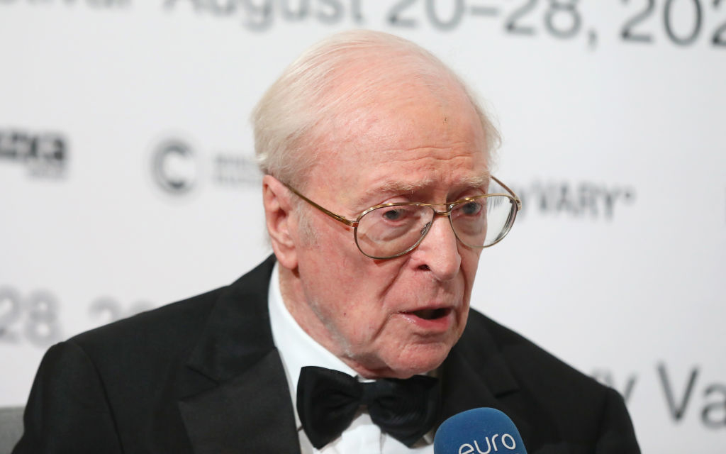 Elderly man in tuxedo being interviewed with event backdrop and microphone