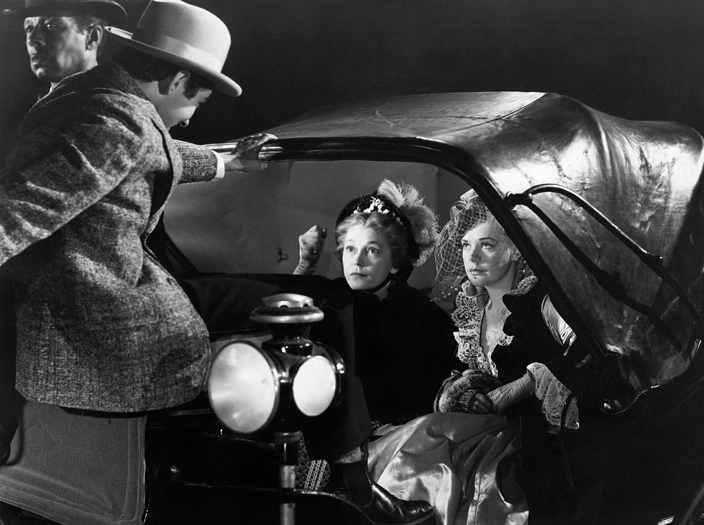 Three vintage film characters, two women seated in an old car, with a man standing outside, all in period attire