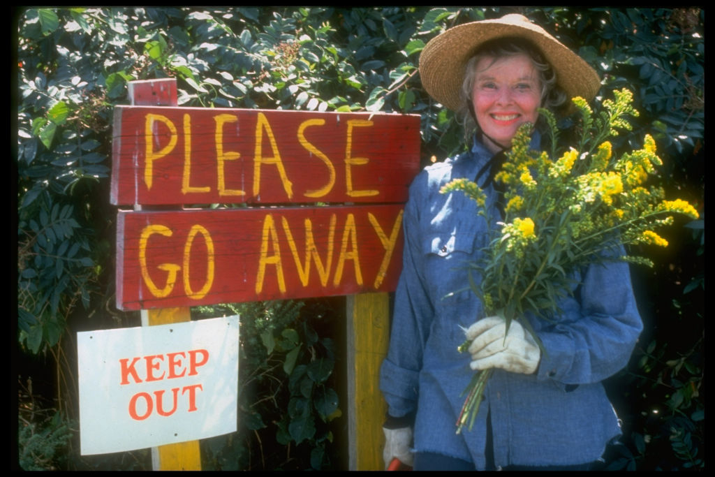 Person in a straw hat holding flowers, standing by signs saying &quot;PLEASE GO AWAY&quot; and &quot;KEEP OUT&quot;