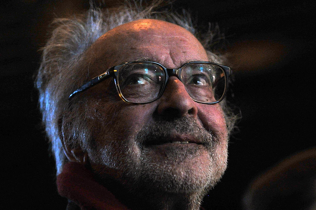 Elderly man with glasses looking up, expression contemplative. He wears a scarf and is in a dimly lit setting