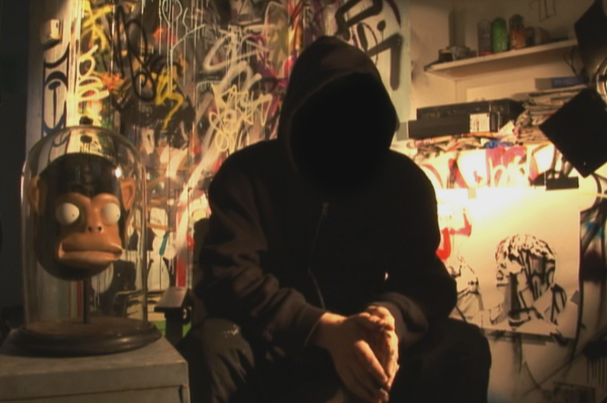 Person in hoodie sitting, anonymous posture, surrounded by urban art and a monkey figure