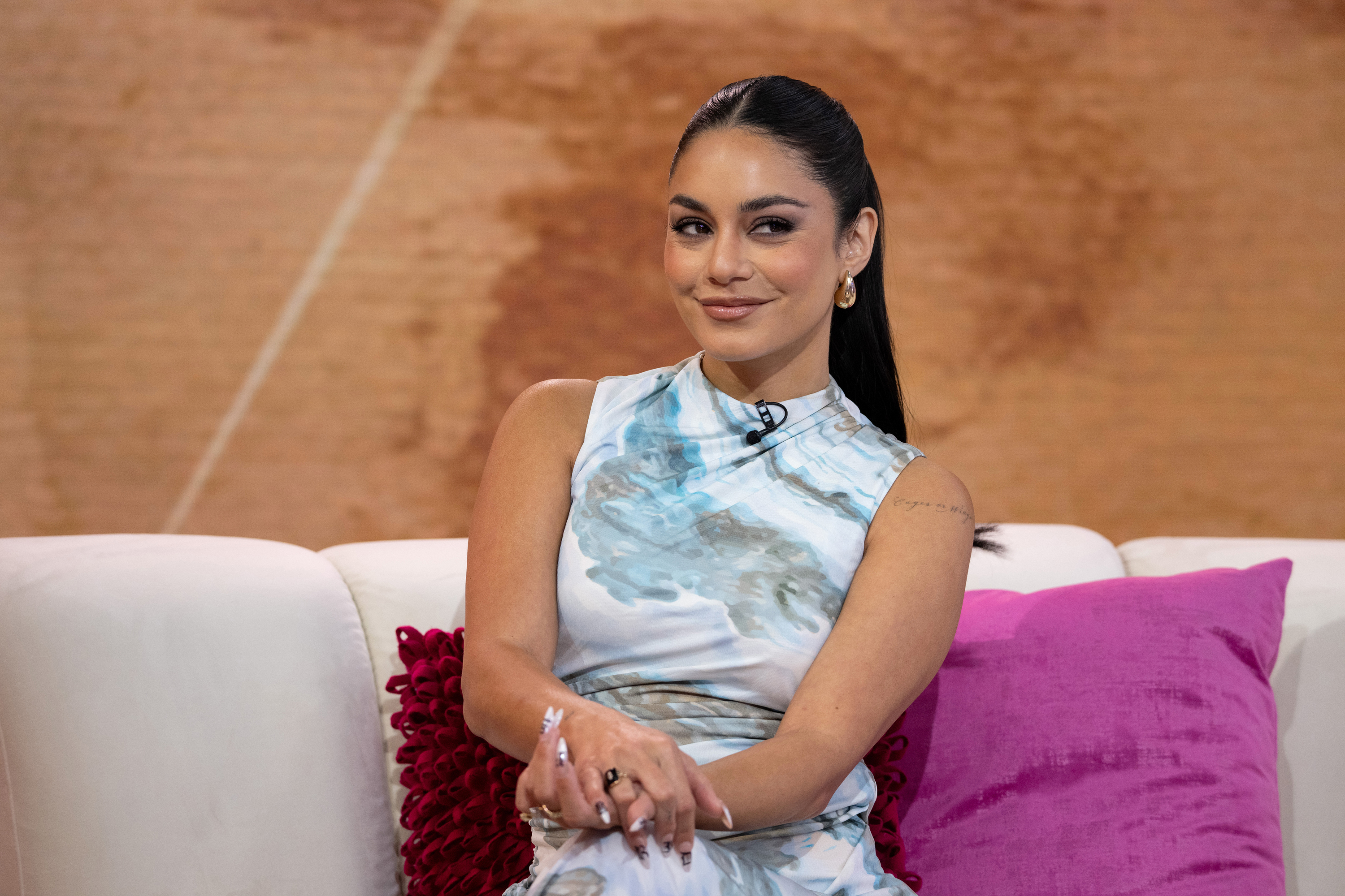 Vanessa Hudgens sits on a sofa wearing a sleeveless patterned dress, posing for a photo