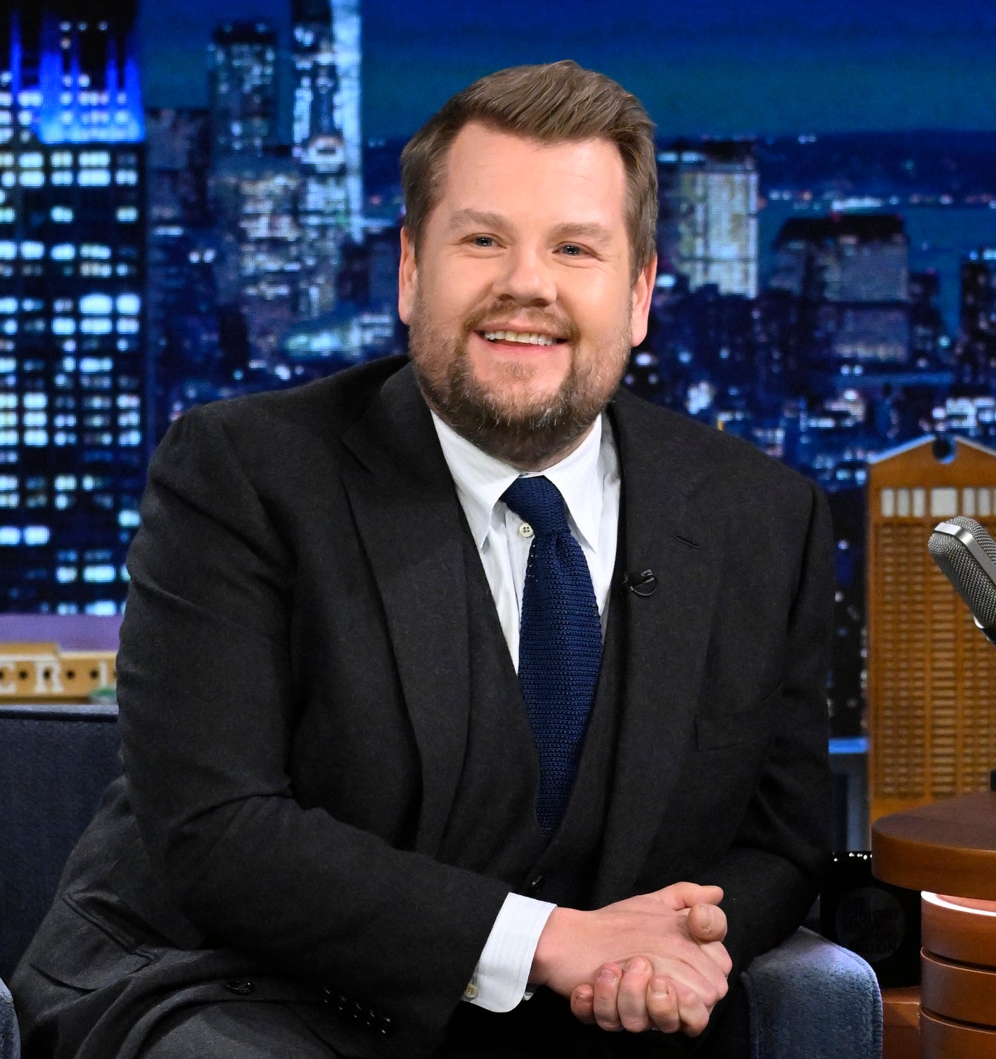 James Corden sitting on a chair in a suit and tie, on a talk show set with cityscape backdrop