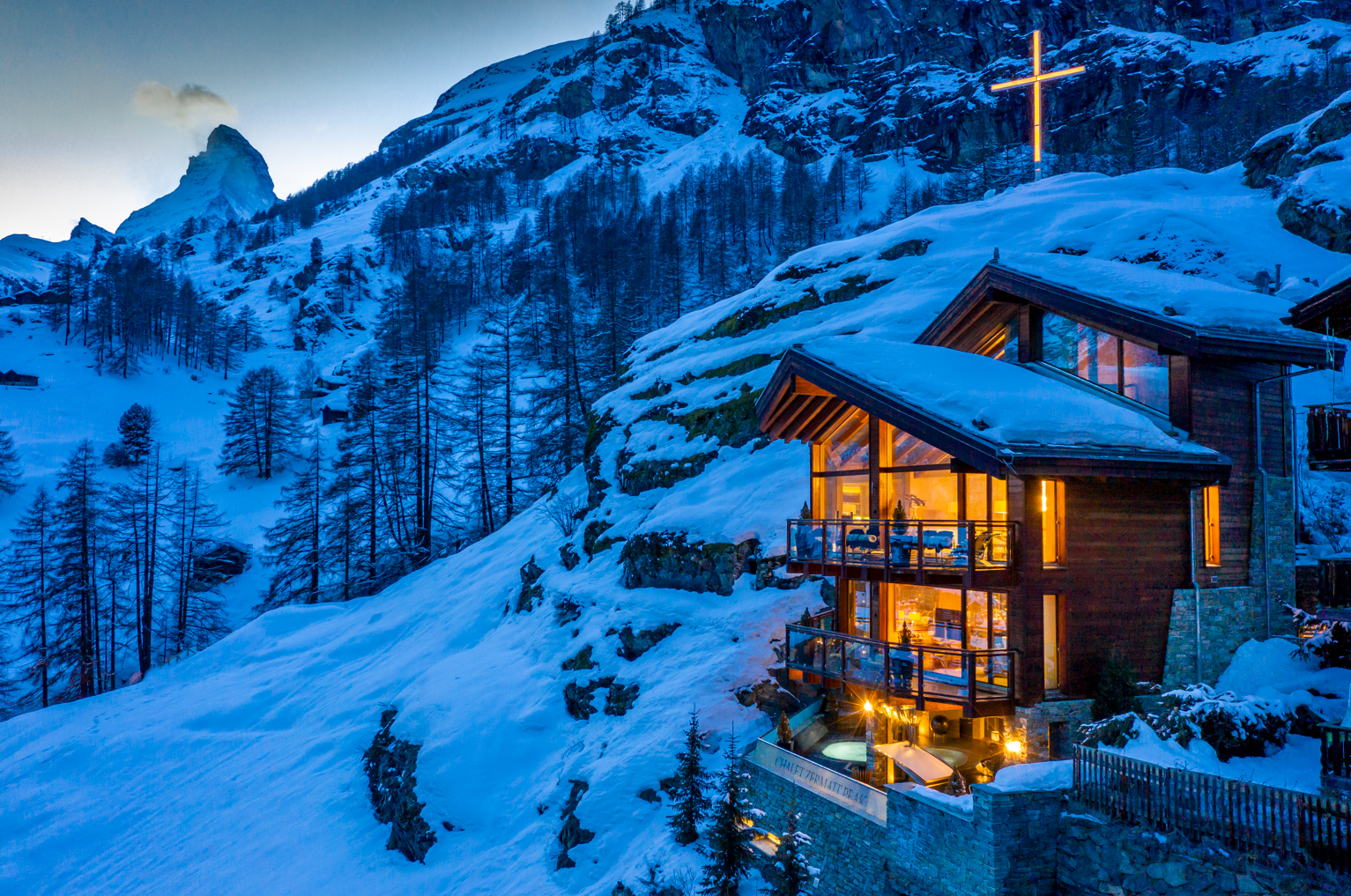 Illuminated chalet in snowy mountains at dusk, with a lit cross on the hillside