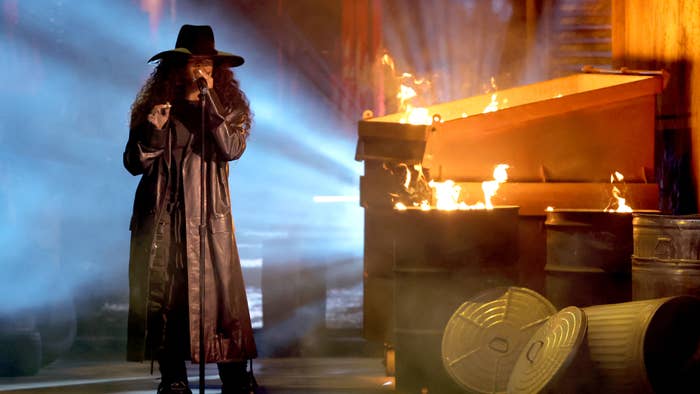 Performer in a long coat and hat singing on stage with a piano on fire in the background