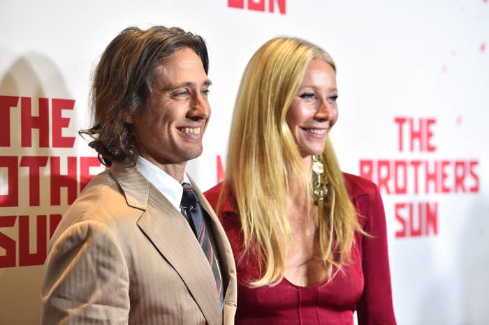 Two individuals smiling at a "The Brothers Sun" event, one in a suit and the other in a v-neck outfit with large earrings