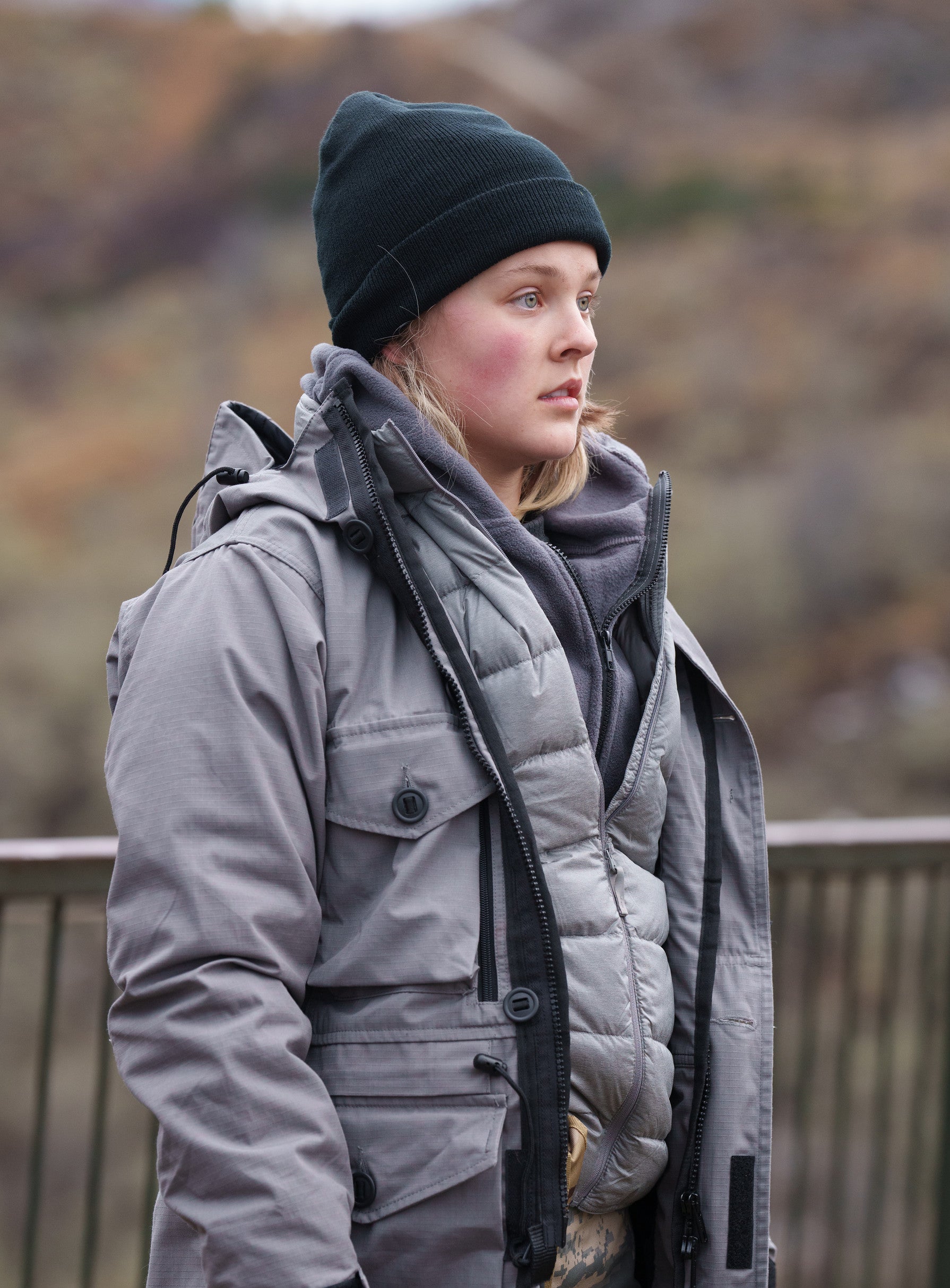 JoJo in warm outdoor layers with a beanie, standing by a railing, looking away thoughtfully
