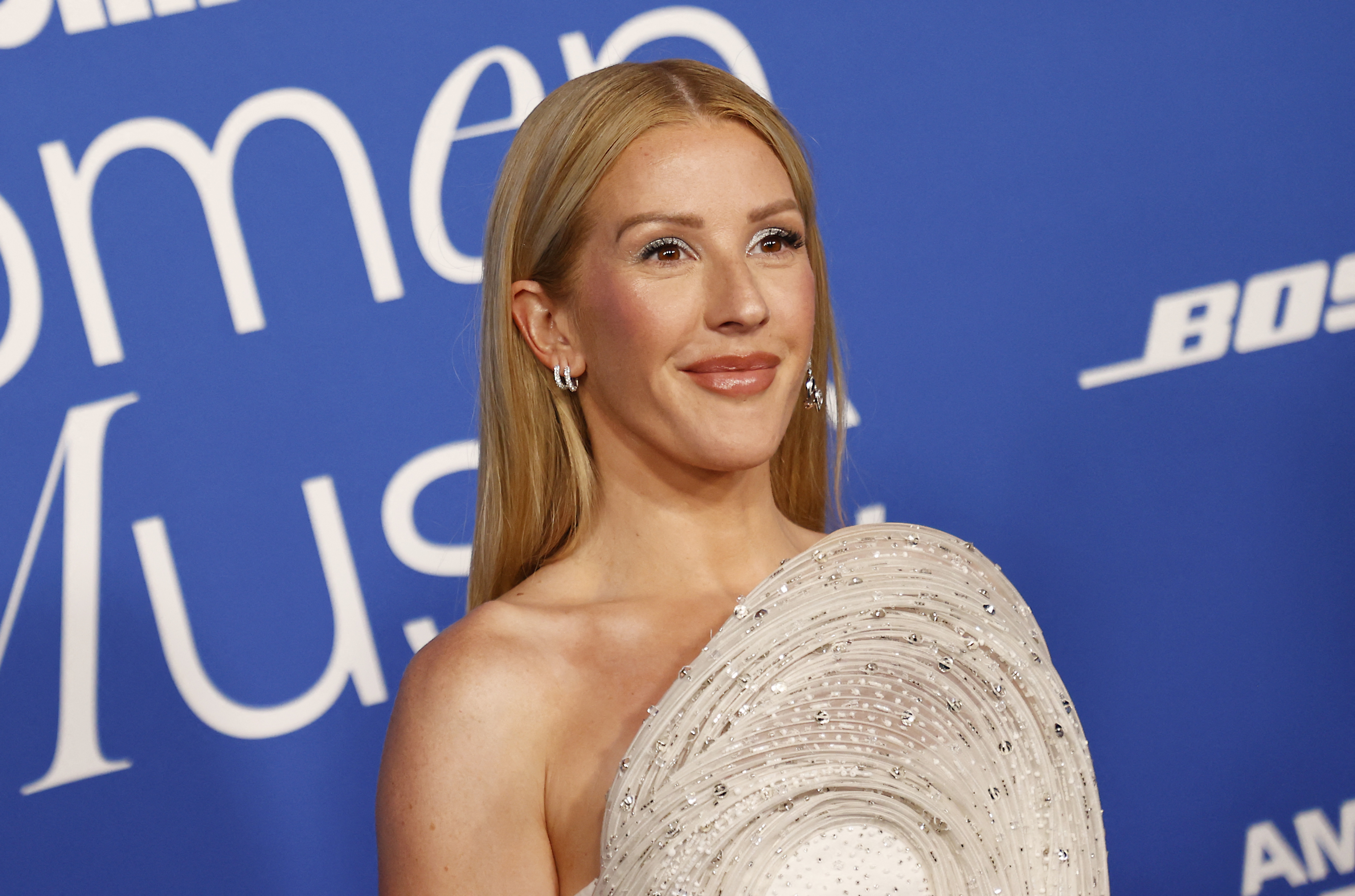 Ellie Goulding smiling, wearing a beaded round-neck gown at a formal event with a logo backdrop