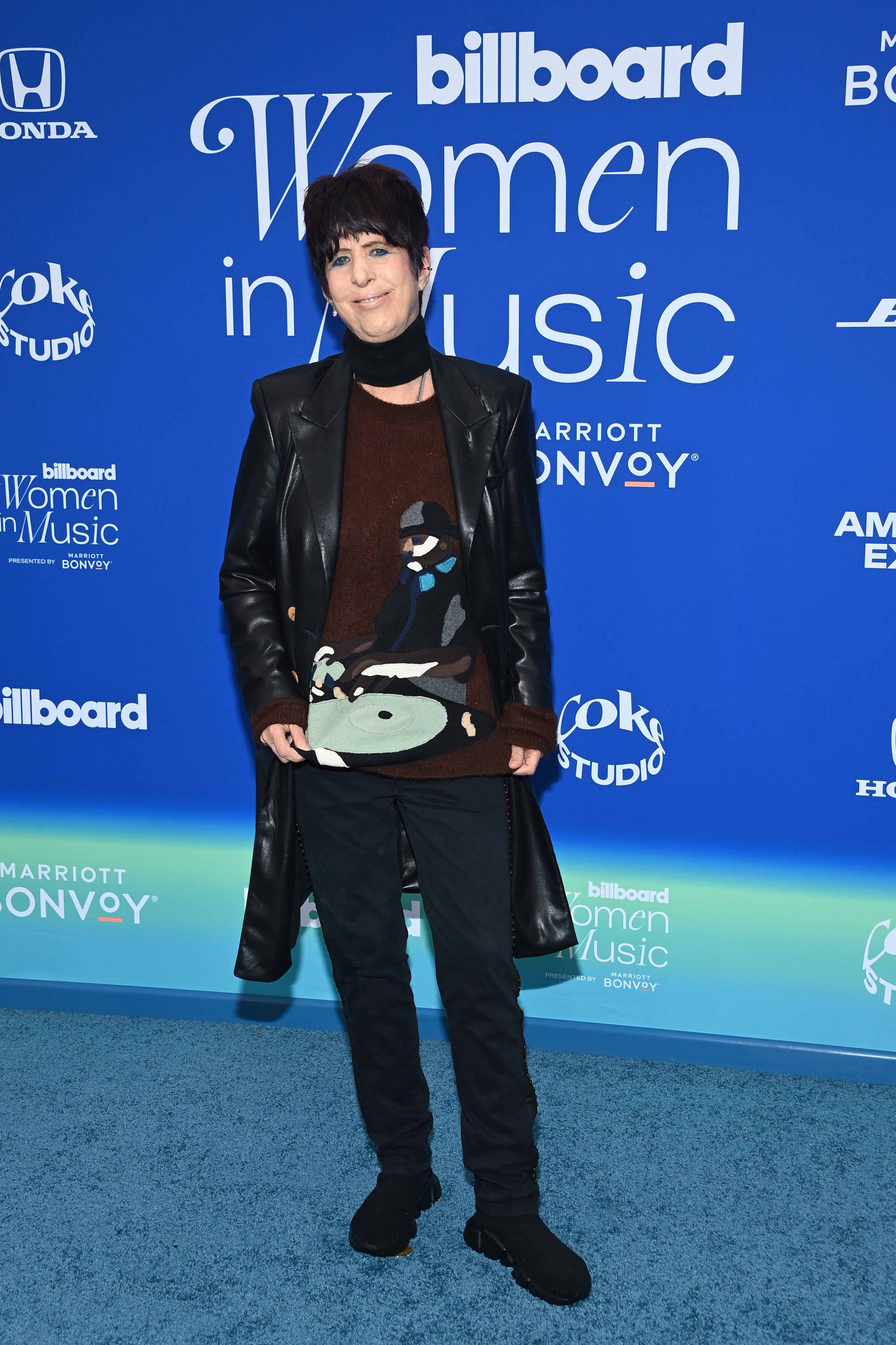 Diane Warren wearing a long leather jacket, pants, chunky boots and a sweater with a DJ design