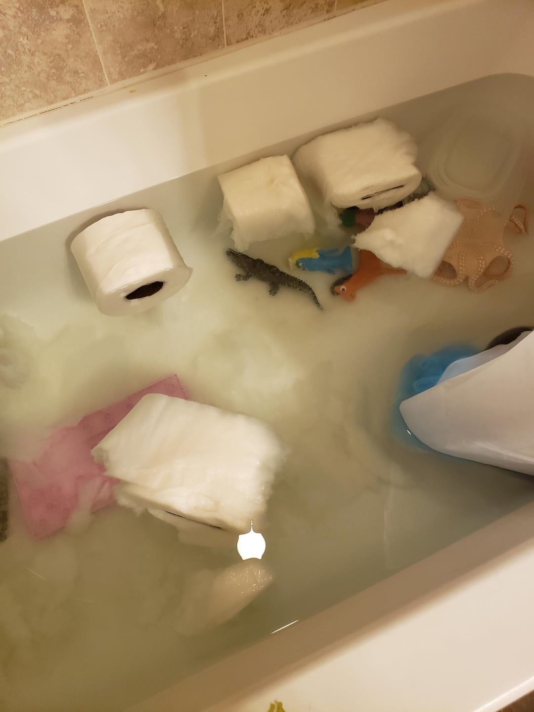 Bathtub filled with water, bubbles, various toys, and a floating roll of toilet paper