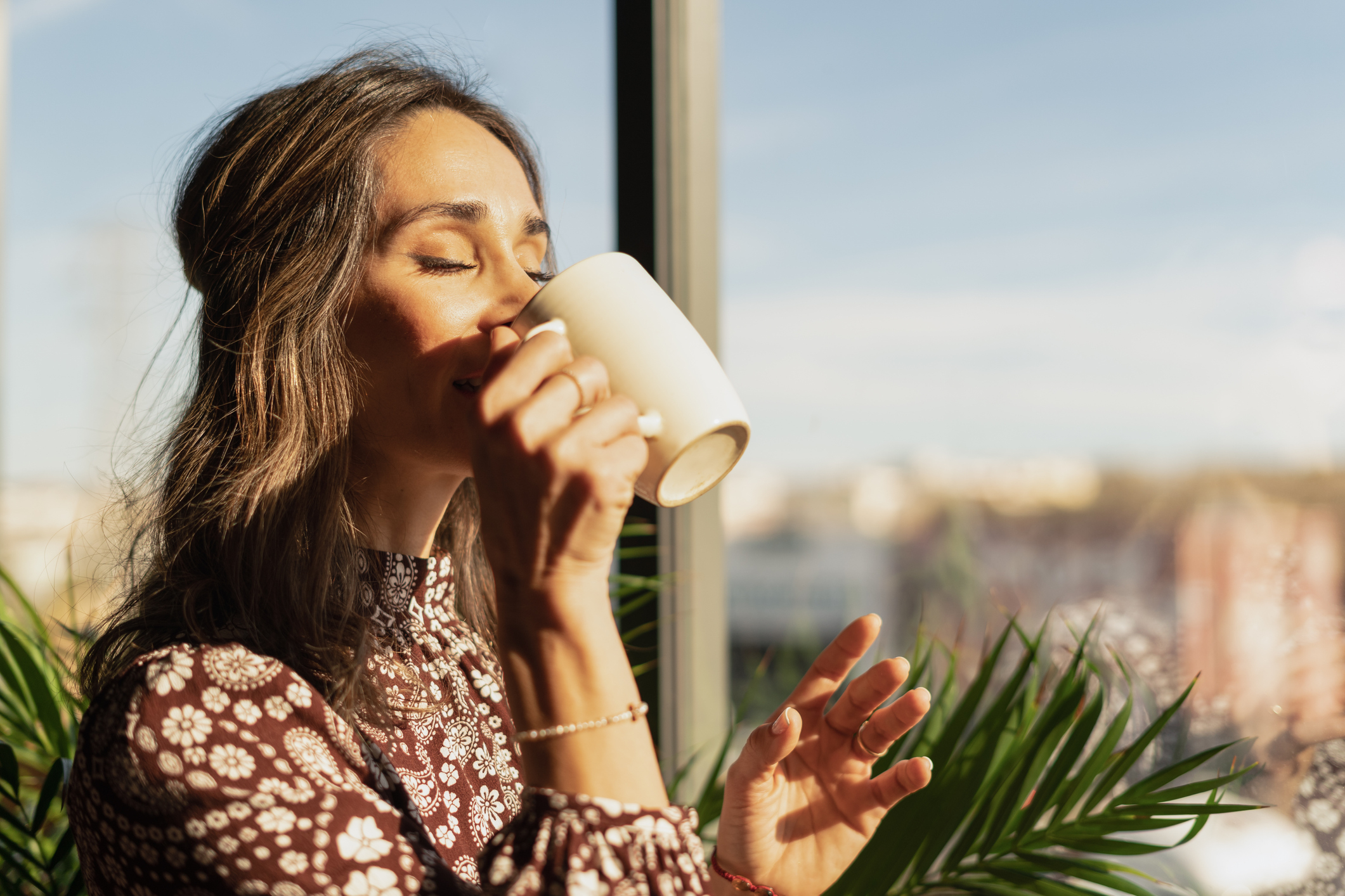 Woman in patterned top sips from a mug by a sunny window