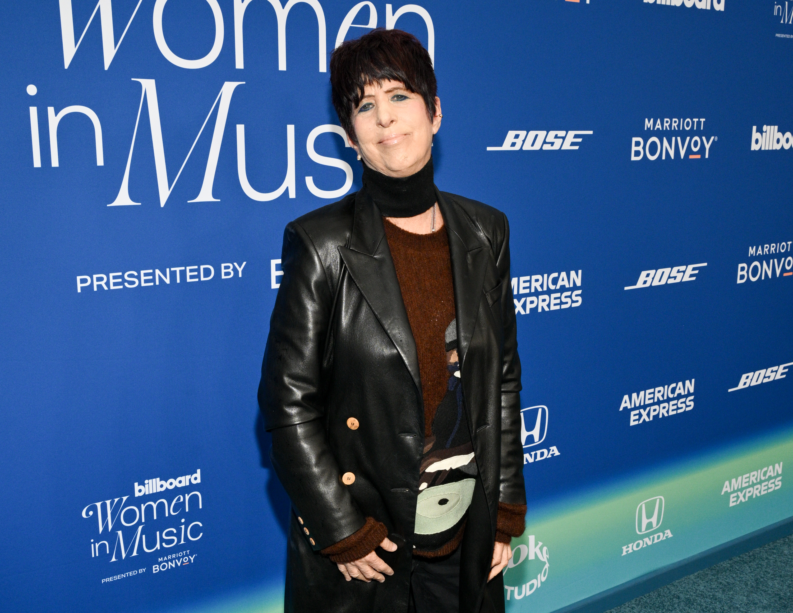Diane Warren at an event wearing a long black coat over a brown top, with a unique white belt accessory