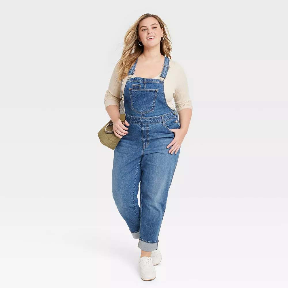 Model in denim overalls and a white long-sleeve top, carrying a clutch, posing for a shopping ad