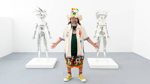 Takashi Murakami stands between two metallic sculptures, wearing a patterned ensemble with eclectic accessories and sneakers