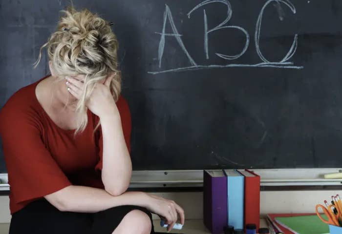 A stressed teacher sitting at a desk with her head in her hands by a blackboard with &quot;ABC&quot; written on it