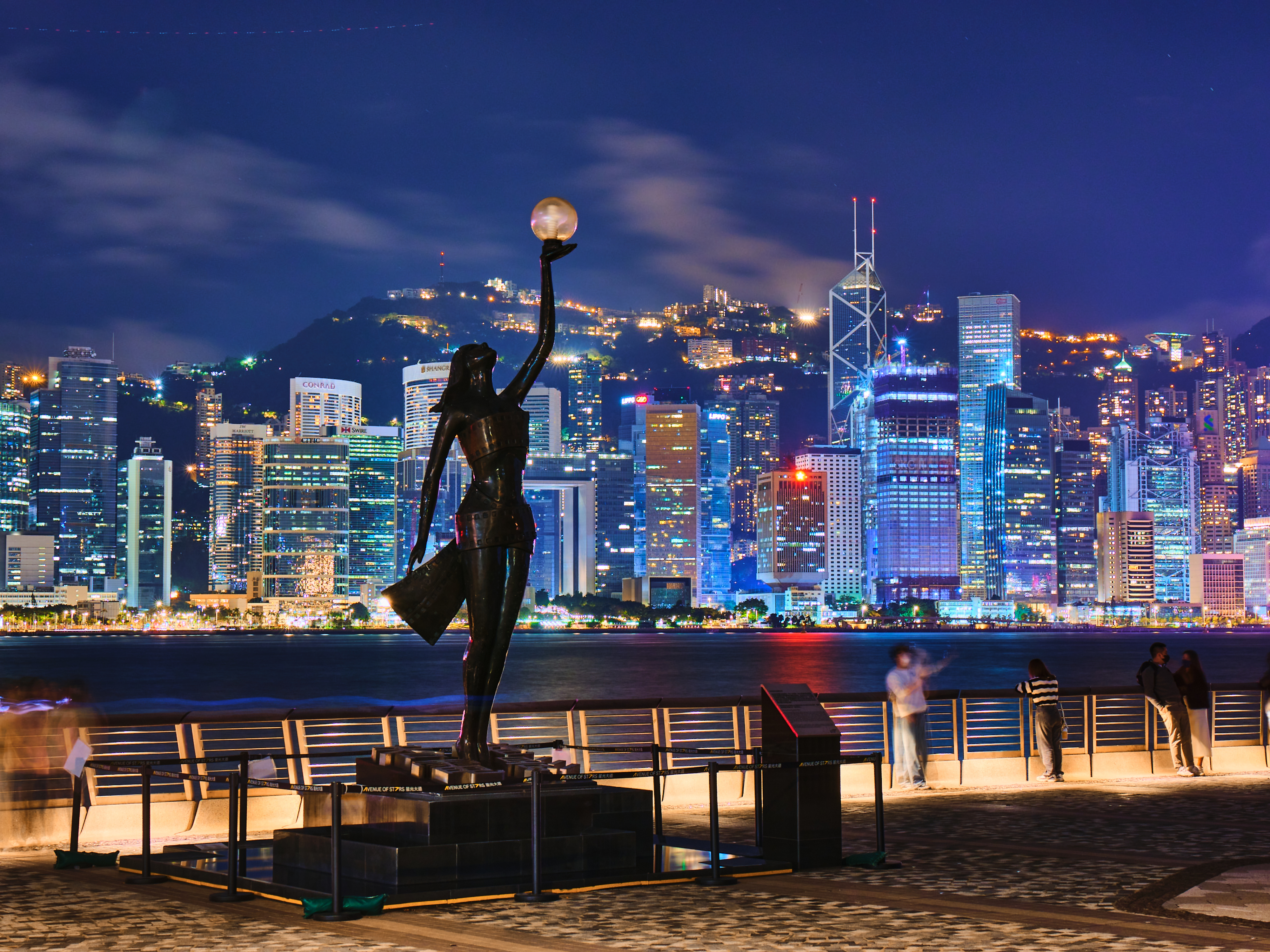 Sculpture of a figure holding aloft a glowing orb by a waterfront with city skyline backdrop at night