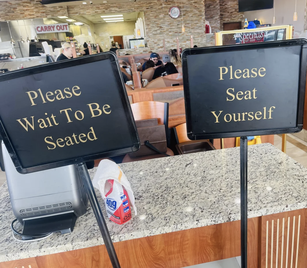 Two contradictory signs at a restaurant: one says "Please Wait to Be Seated" and the other "Please Seat Yourself."