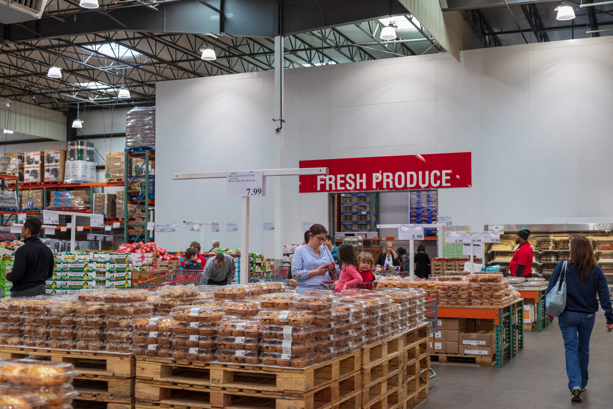 Shoppers at a warehouse store with &quot;FRESH PRODUCE&quot; sign, browsing items on pallets