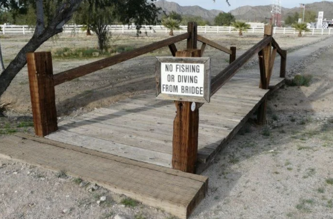 Wooden, small footbridge above dry dirt with a sign that reads "NO FISHING OR DIVING FROM BRIDGE"