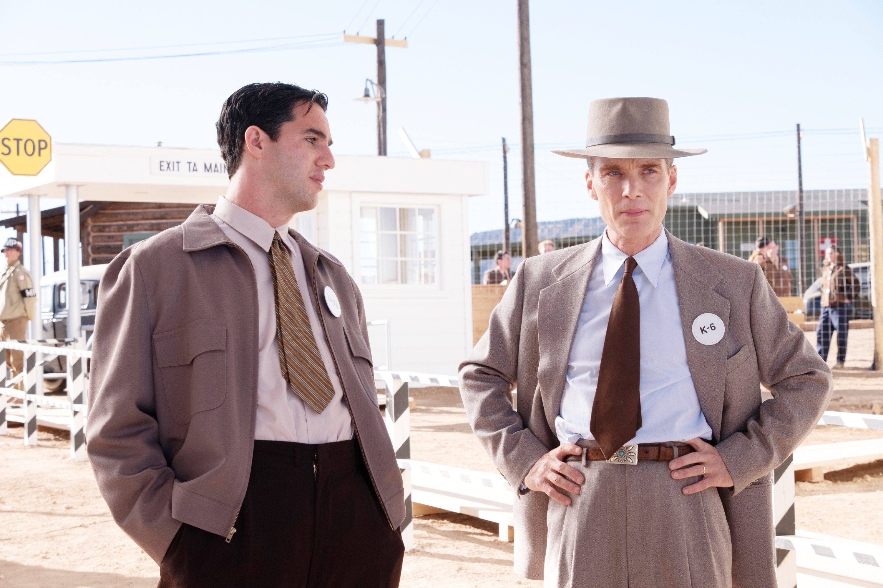 Cillian Murphy as Oppenheimer stands next to Benny Safdie as Edward Teller in an outdoor setting with a &quot;STOP&quot; sign and buildings behind them