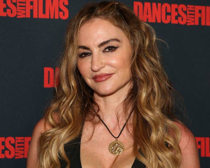 drea de matteo at event, styled in a dark v-neck outfit with a medallion necklace, posing with a slight smile