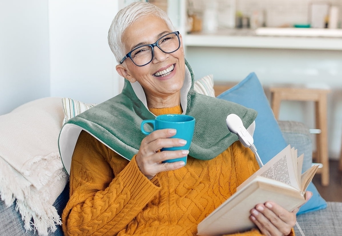 Person smiling, holding a book and a mug, wearing glasses and a cable knit sweater, sitting with pillows