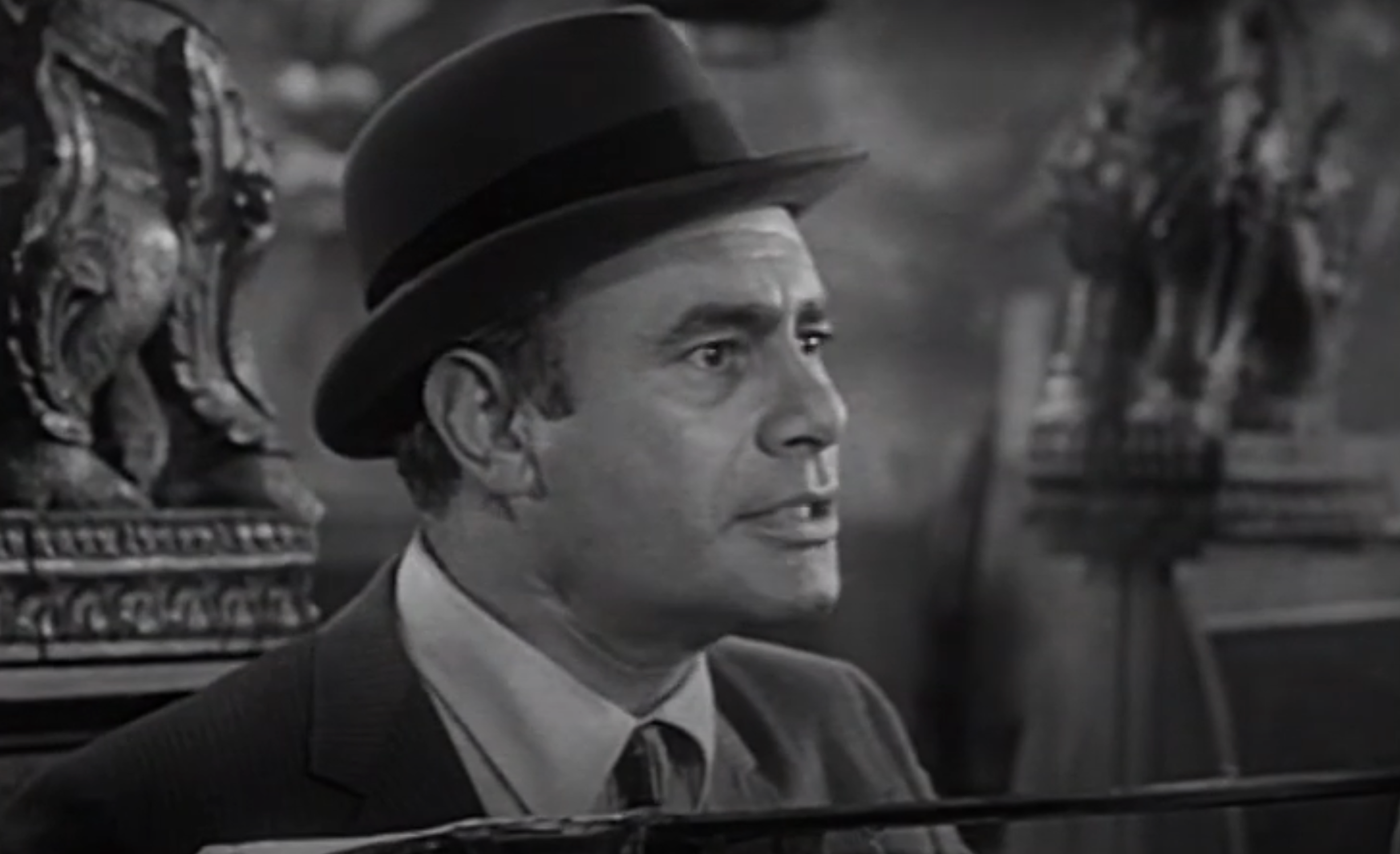 Man in a fedora and suit, looking to the side, with a concerned expression in a black-and-white film scene