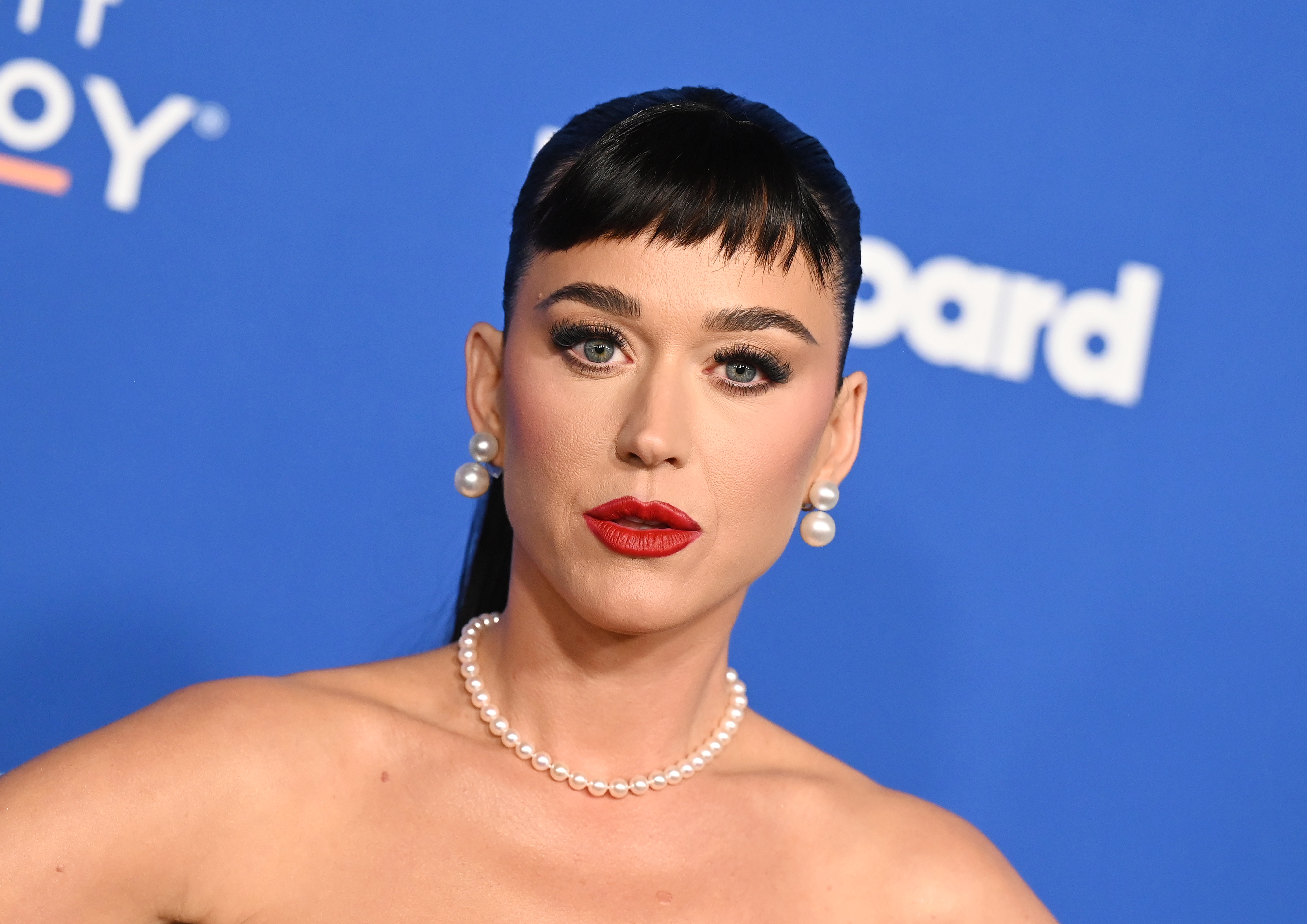 Katy posing on at a media event, wearing a pearl necklace and earrings, with bold lipstick