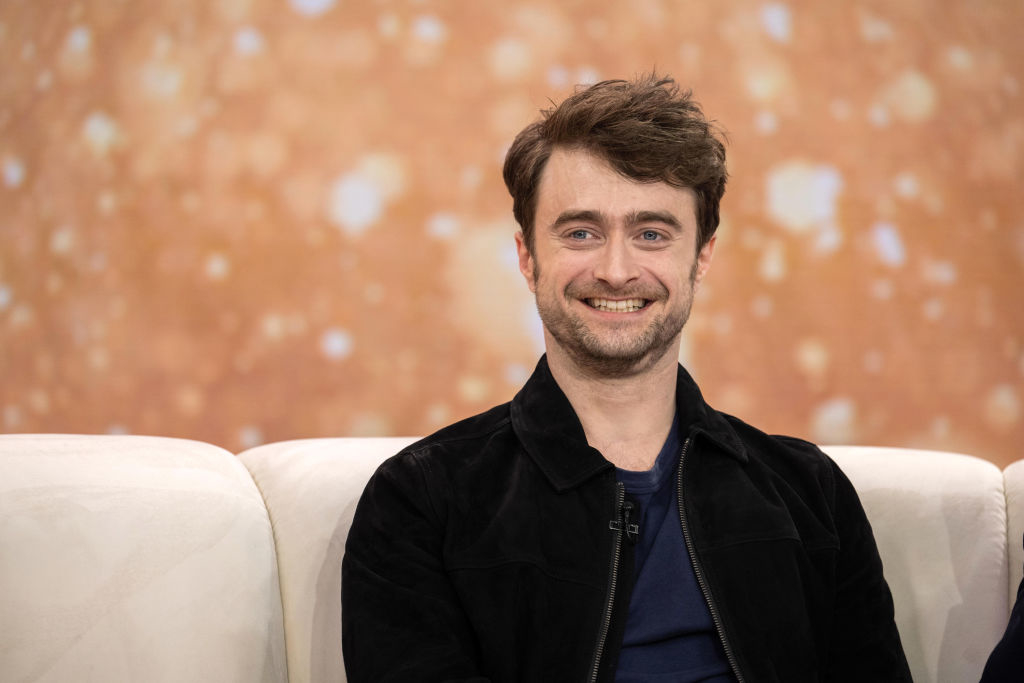 Daniel Radcliffe sitting, smiling in a black jacket and blue shirt, on a white couch