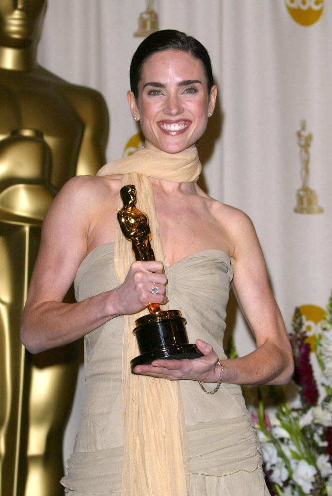Jennifer Connelly smiling and holding her Oscar, wearing a strapless pale dress with a scarf
