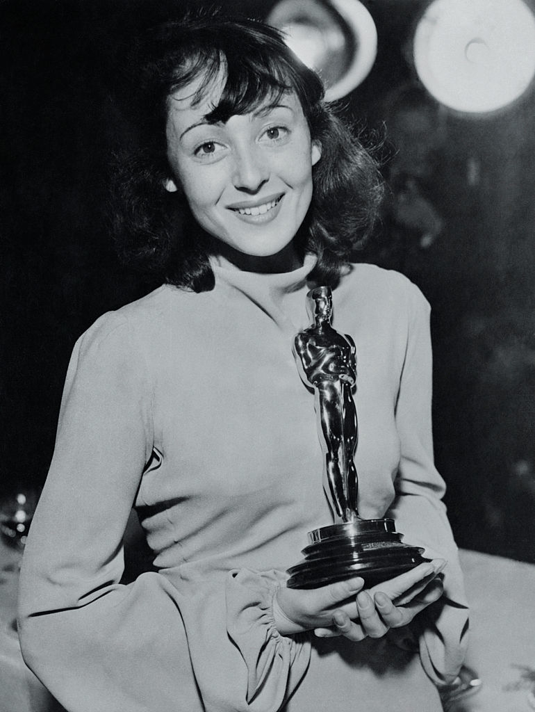 Luise in a button-up top holds an Oscar trophy, smiling