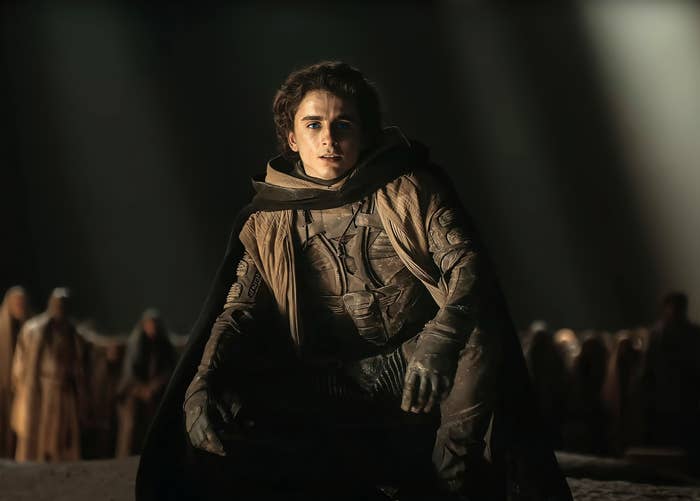 Paul Atreides in a textured coverall with a cloak stands center in a dramatic scene from the film &quot;Dune&quot;