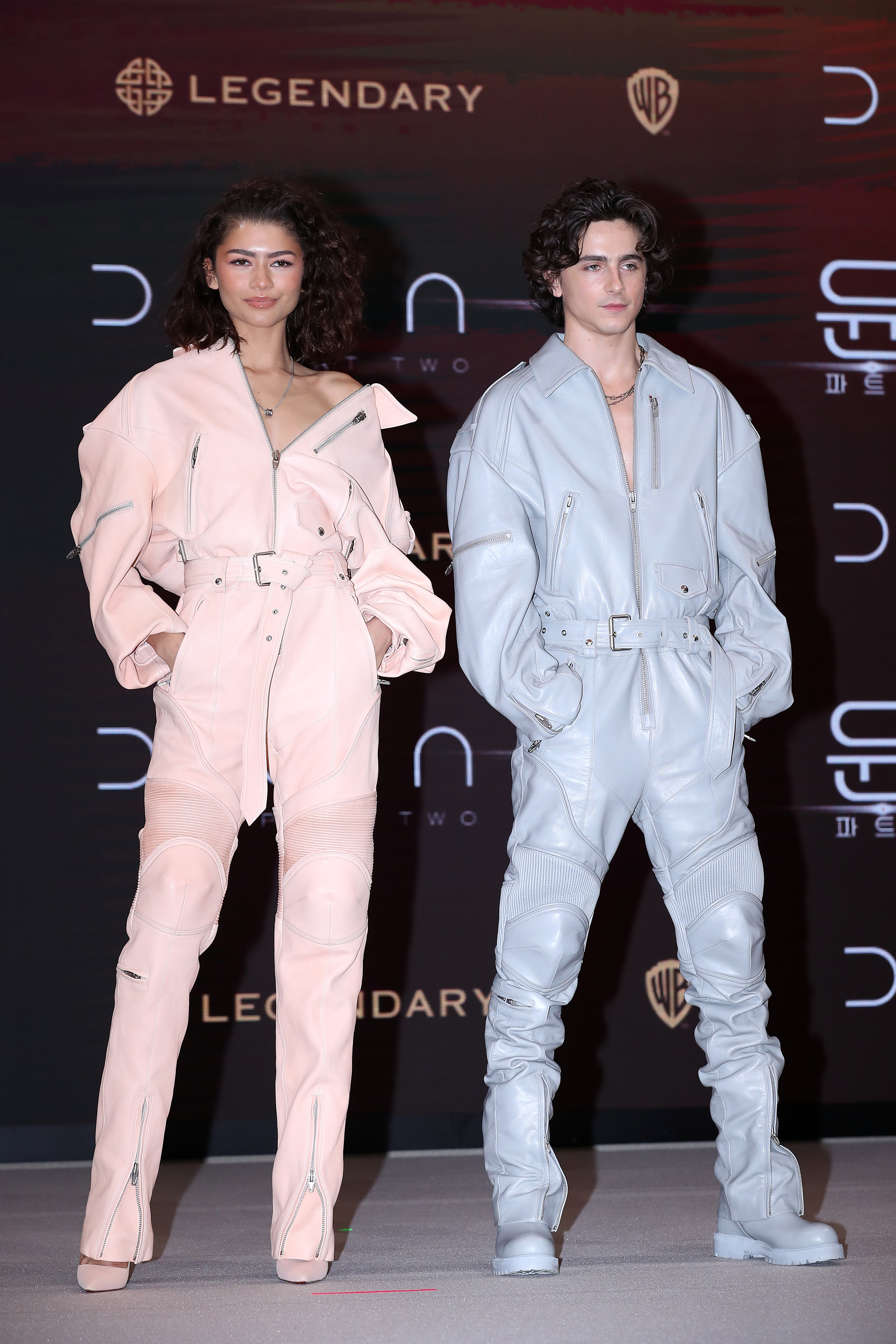Zendaya and Timothée Chalamet stand side by side; both in stylish belted jumpsuits and boots