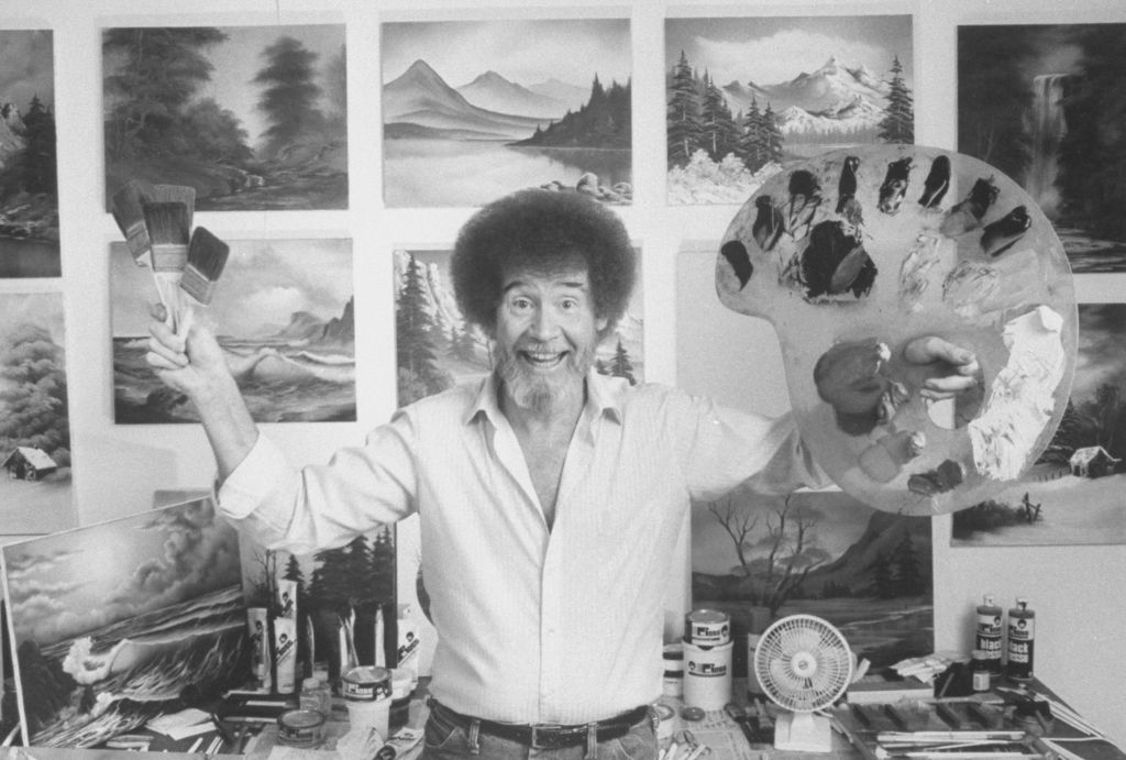 Man with afro smiling, holding a paint palette, surrounded by landscape paintings
