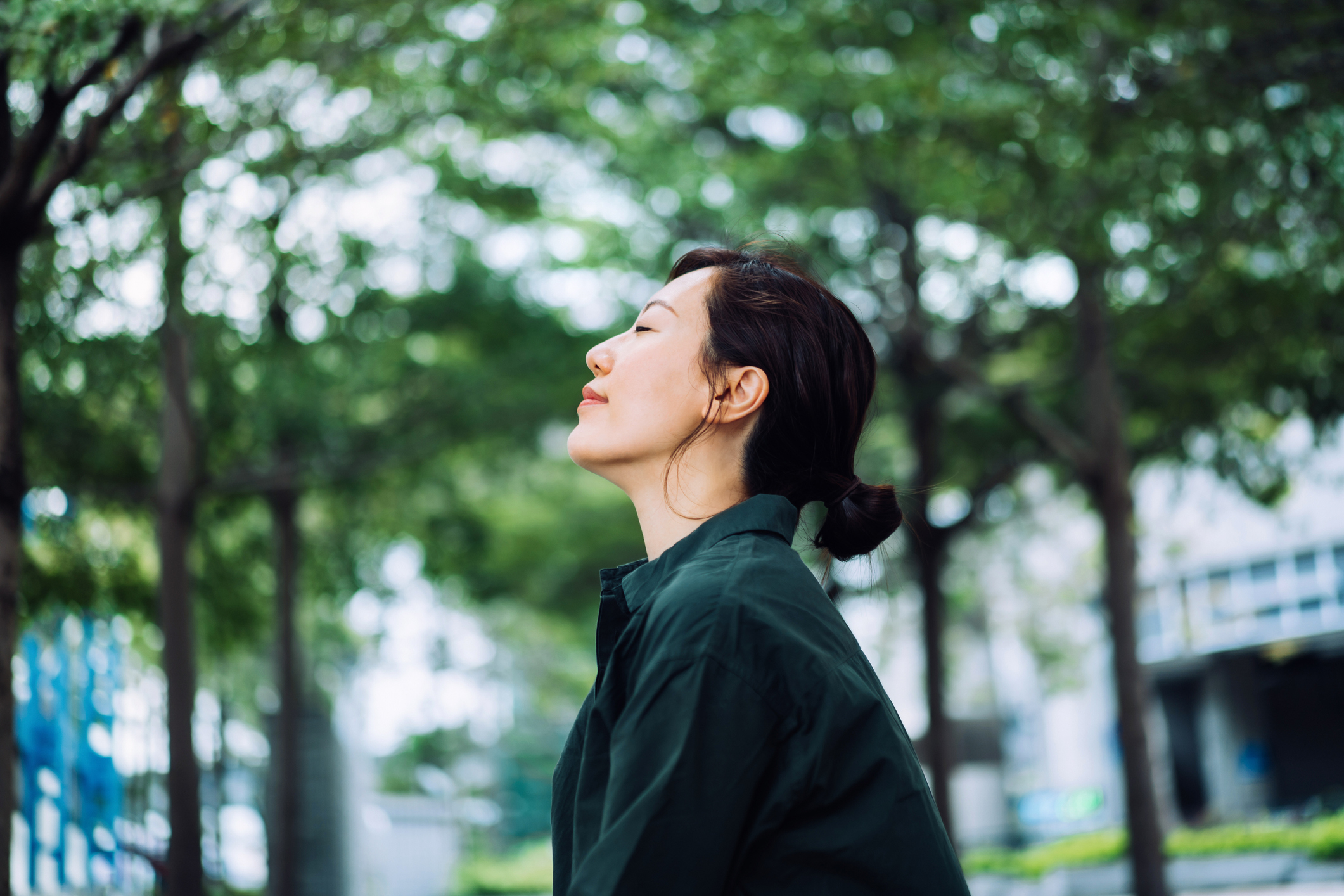 Woman in casual wear with eyes closed and head raised, enjoying fresh air among trees in an urban setting