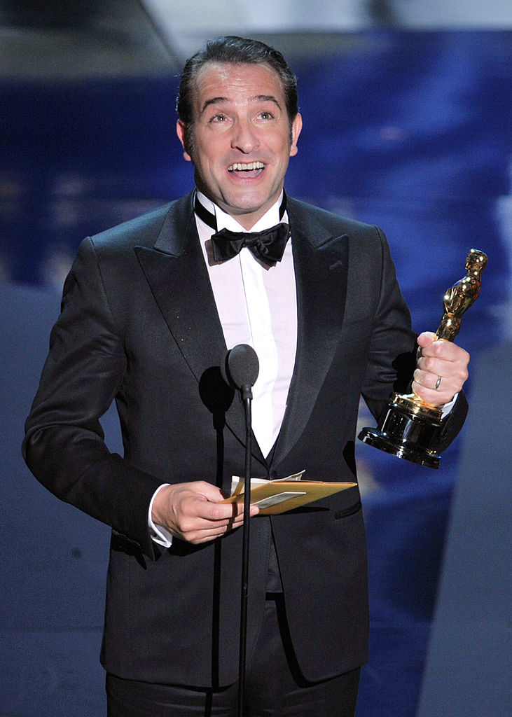 Jean, in a tuxedo, holding his Oscar statuette before a microphone, expressing joy
