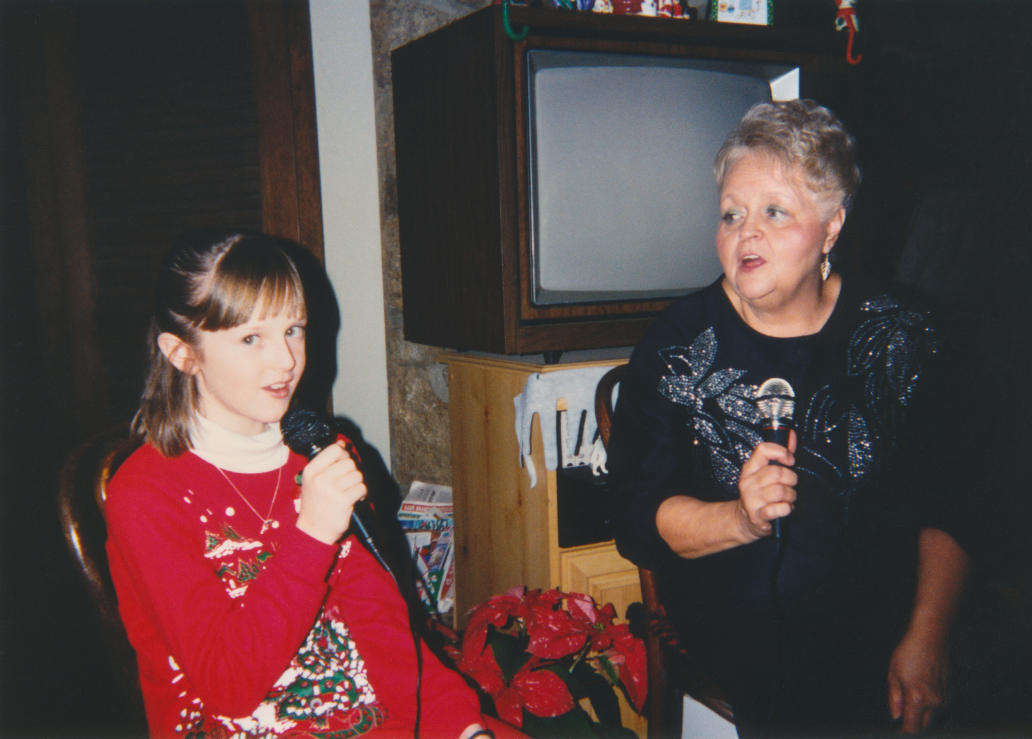 Girl and older woman singing into microphones, casual home setting with an old-fashioned cathode ray TV in the background