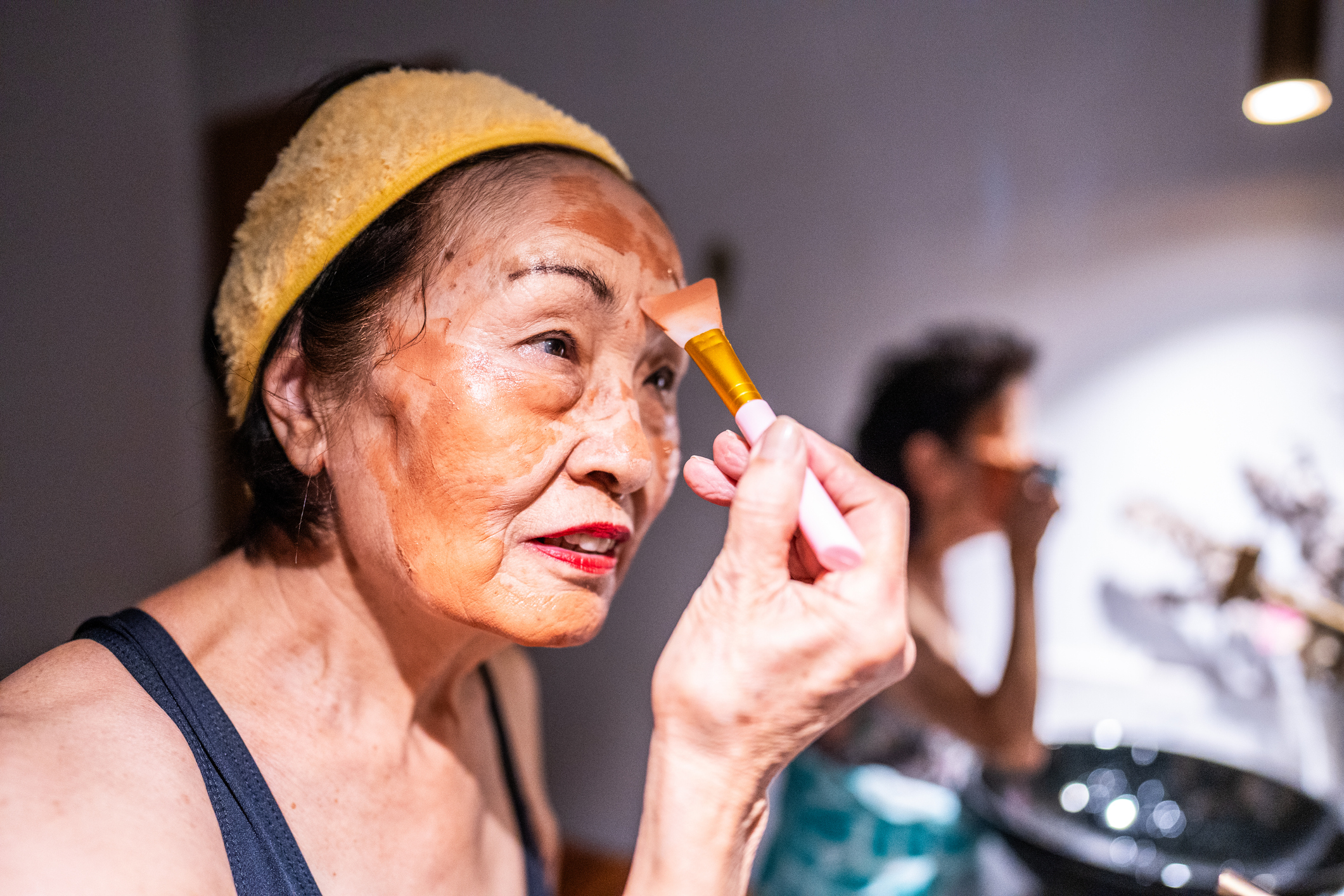 Older woman applying makeup with a brush, standing in front of a mirror