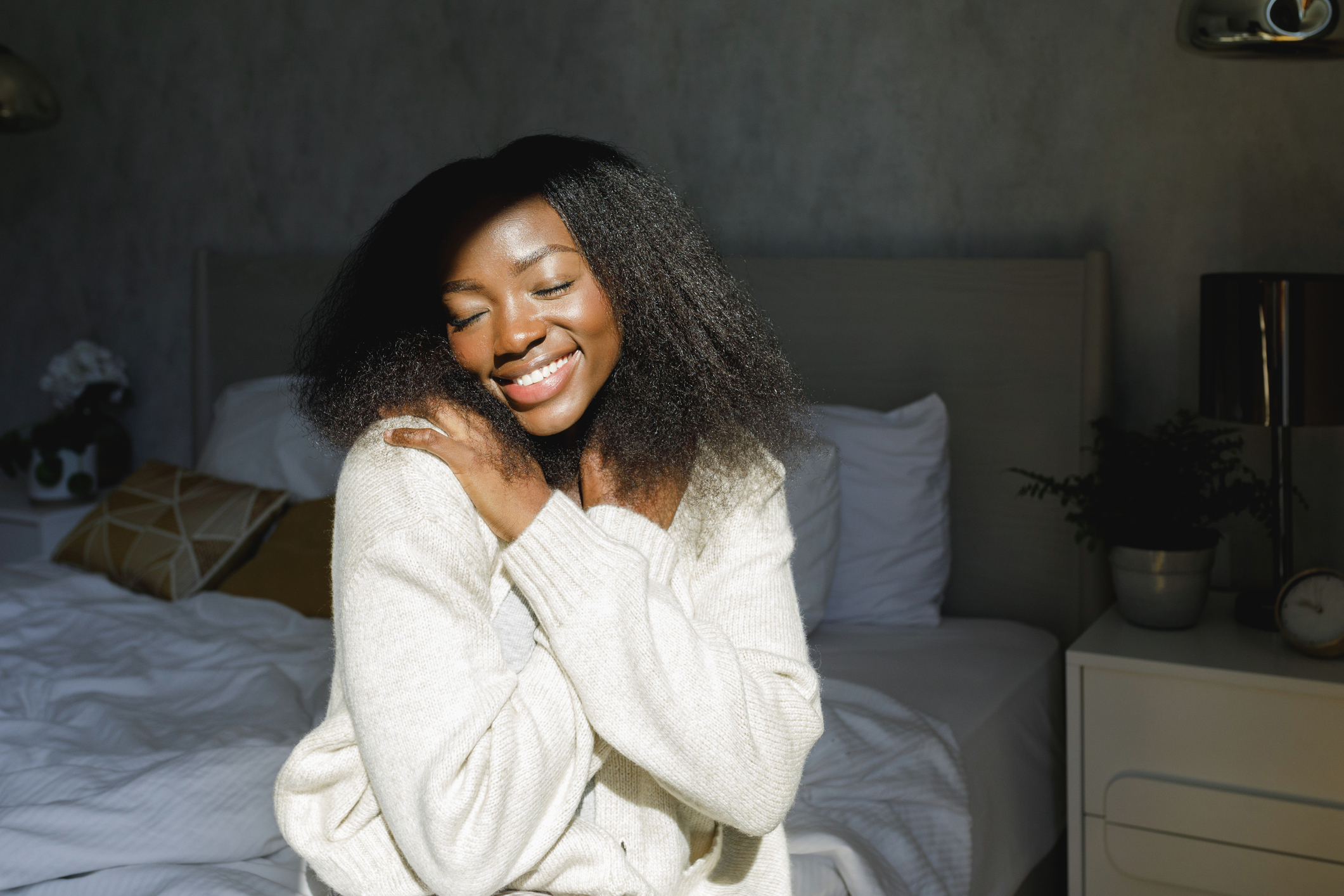 A woman in a knit sweater smiling with eyes closed, embracing herself in cozy bedroom setting