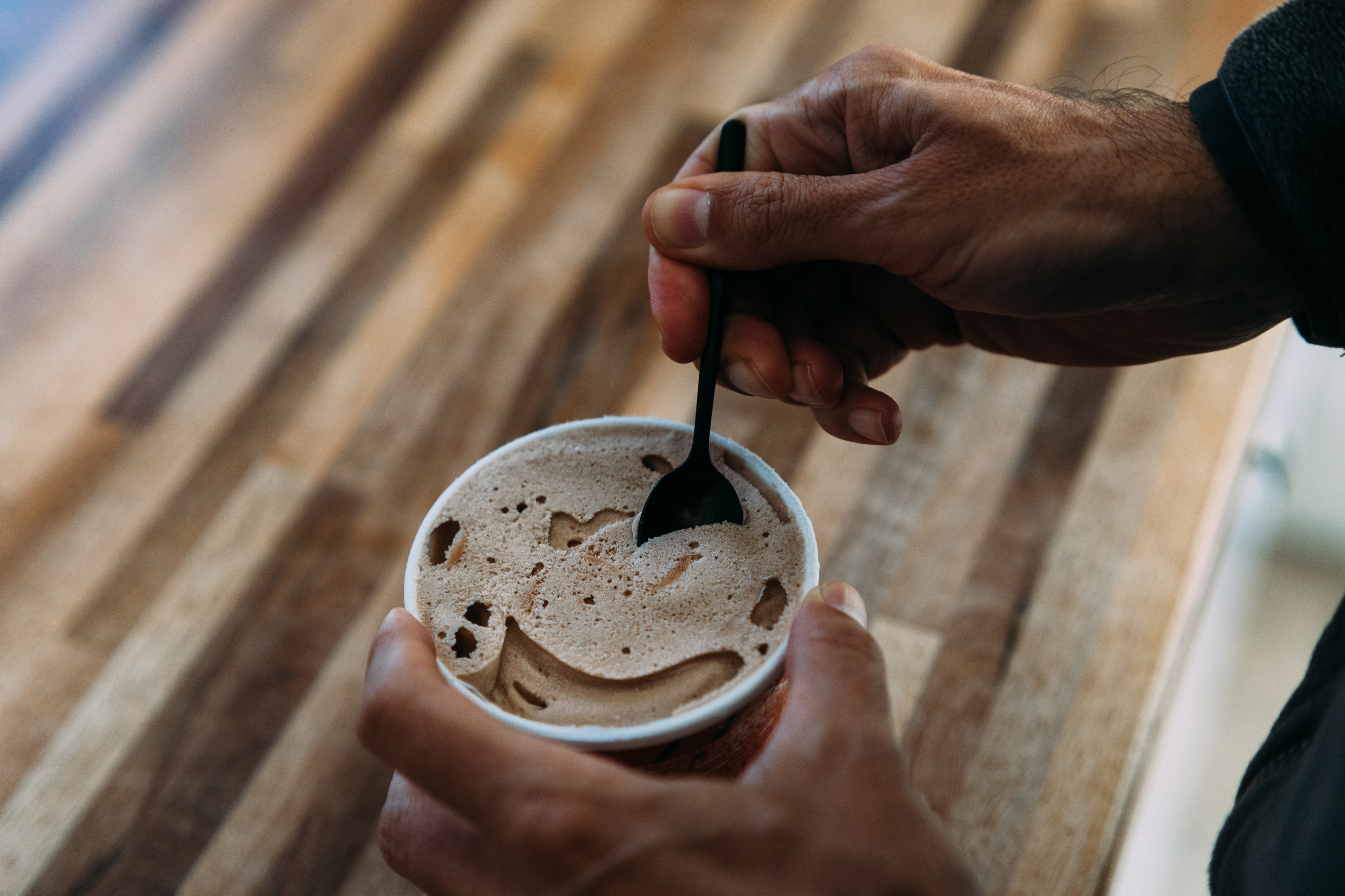 Person holding a cup of frothy coffee with a spoon, on a wooden surface