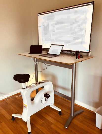 A standing desk with a laptop, tablet, monitor, and an under-desk exercise bike