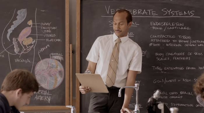 Teacher with clipboard in front of a chalkboard labeled &quot;Vertebrate Systems&quot; in a classroom scene