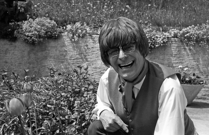 Man in vest and tie, smiling, with tousled hair, standing in a garden