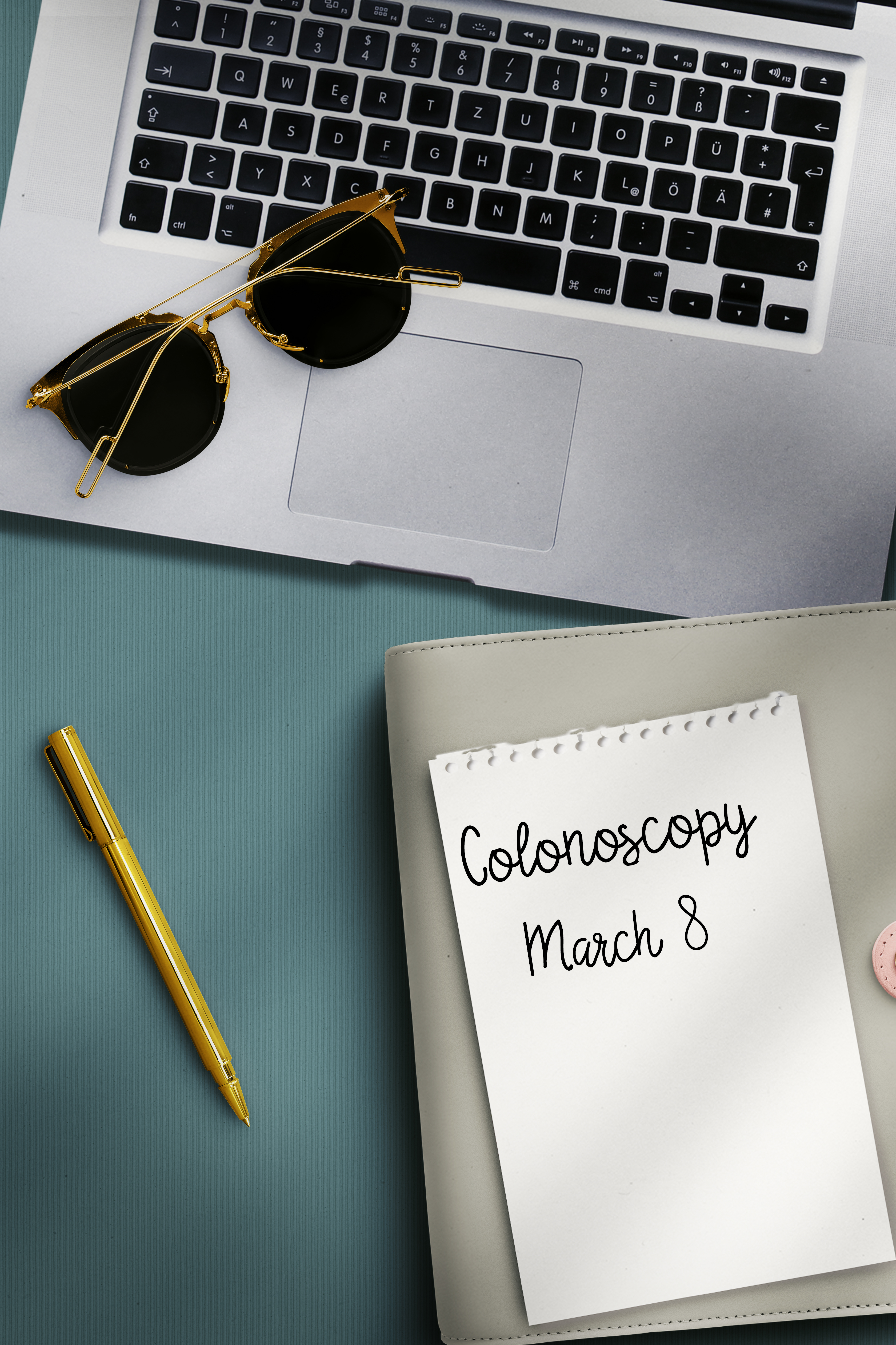 Notepad with handwritten &#x27;Colonoscopy March 8&#x27; beside laptop, sunglasses, and pen