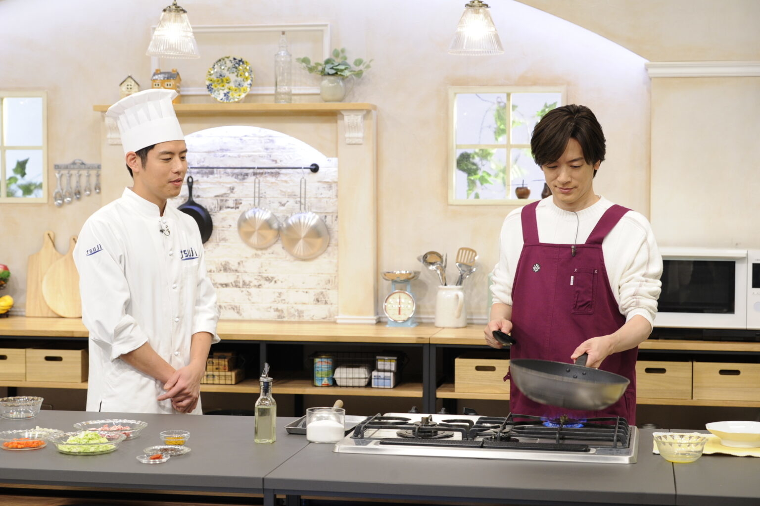 二人の男性がキッチンで料理をしている。一人はシェフの制服を、もう一人はエプロンを着用している。